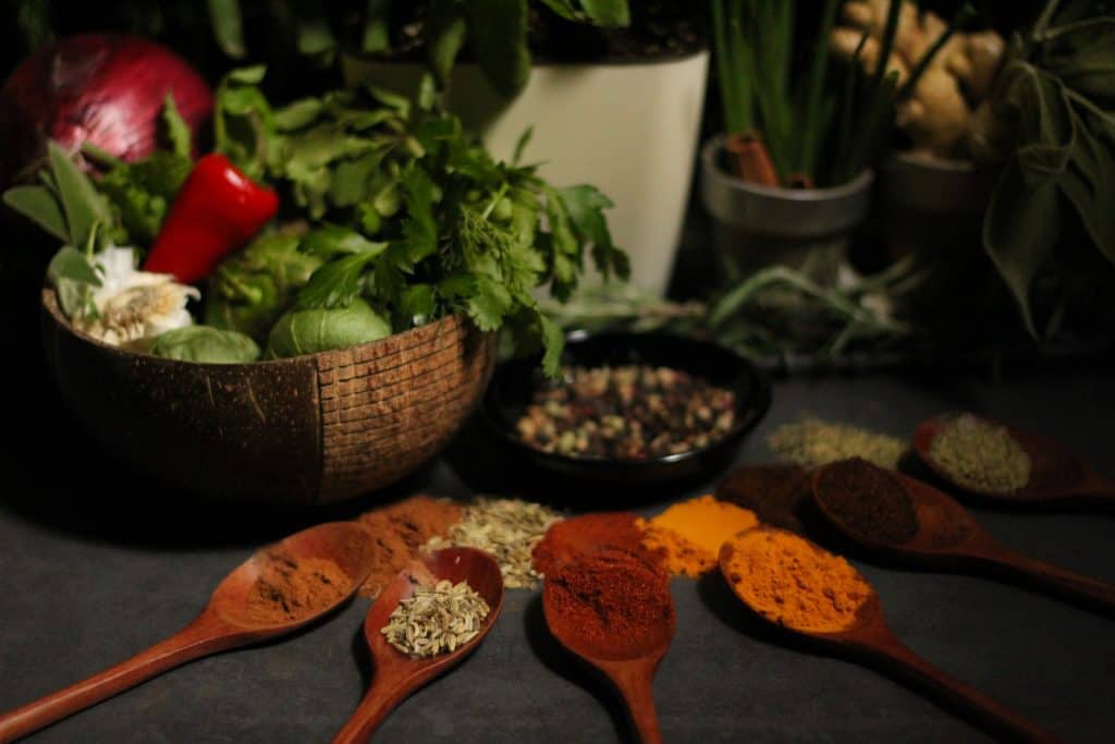 Heavenly Spiced dark and moody display of herbs in brown bowl and in background with colorful spices on brown wooden spoons