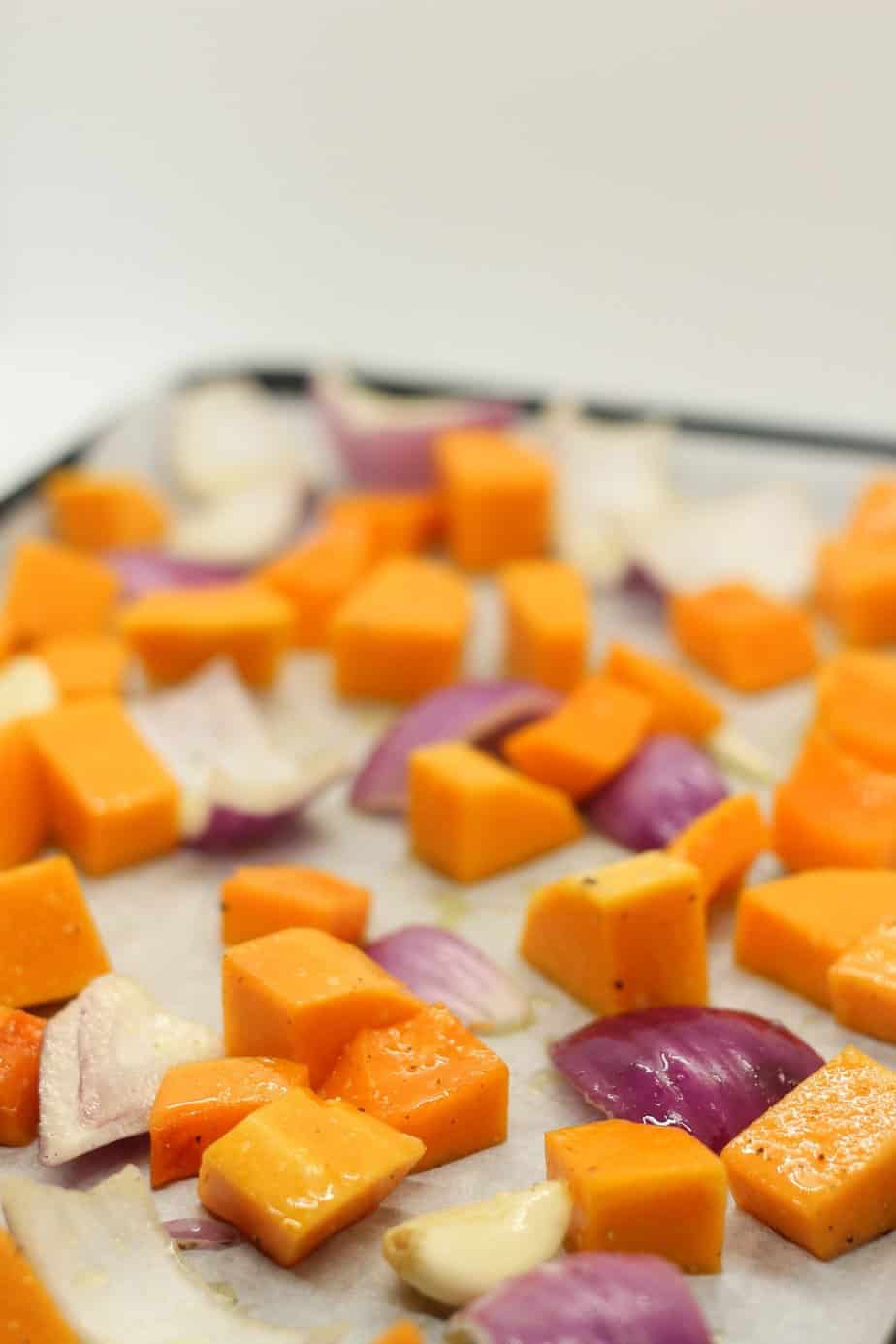 Diced butternut squash, red onion, and peeled garlic on a sheet baking pan lined with parchment.