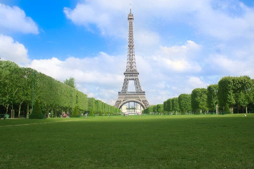 Eiffel tower at the end of a large green field lined with trees.