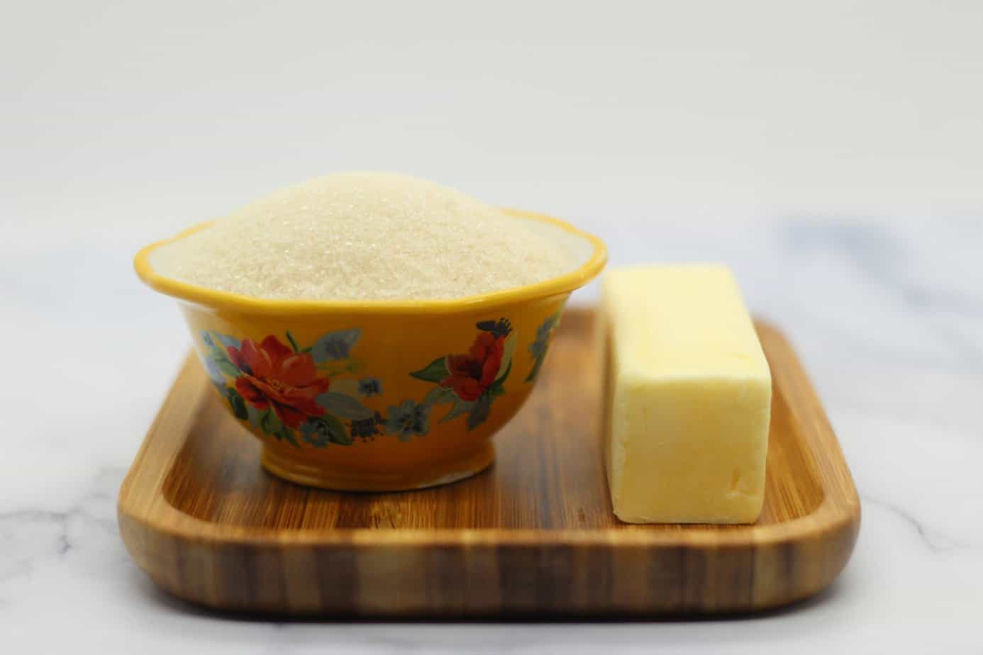 sugar in a yellow floral bowl and stick of butter on a light brown wooden plate on a white marble surface