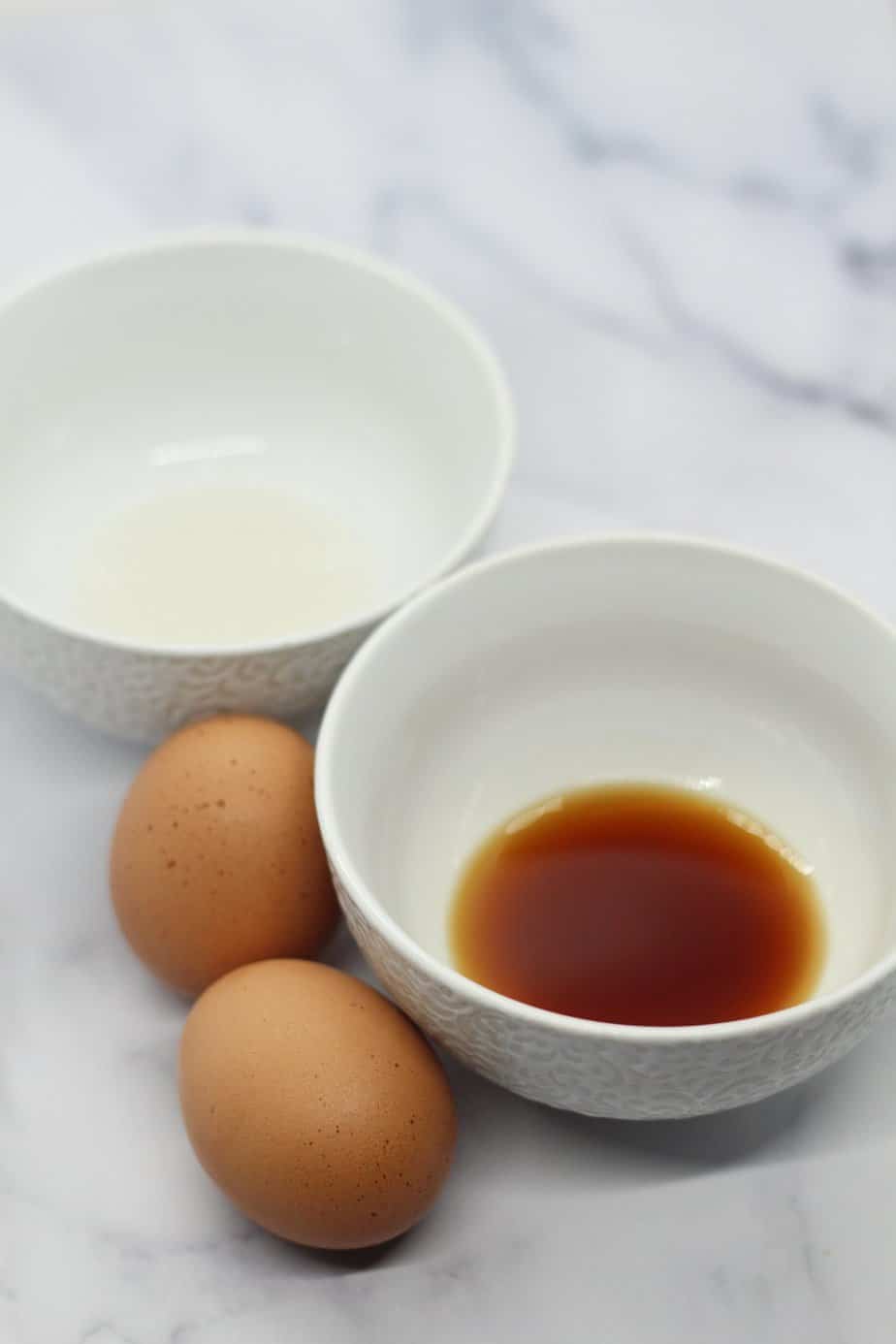 two brown eggs and two separate white bowls of vanilla extract and almond extract on white marble surface