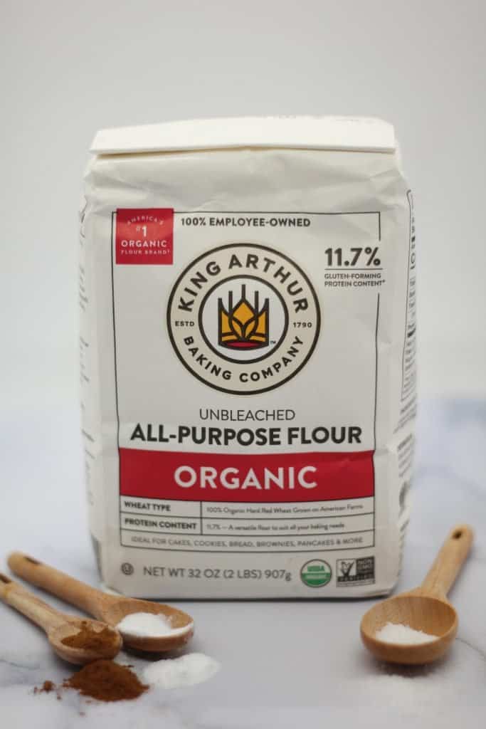 bag of organic King Arthur all-purpose flour next to wooden spoons filled with salt, baking soda, baking powder, and cinnamon on a white marble surface