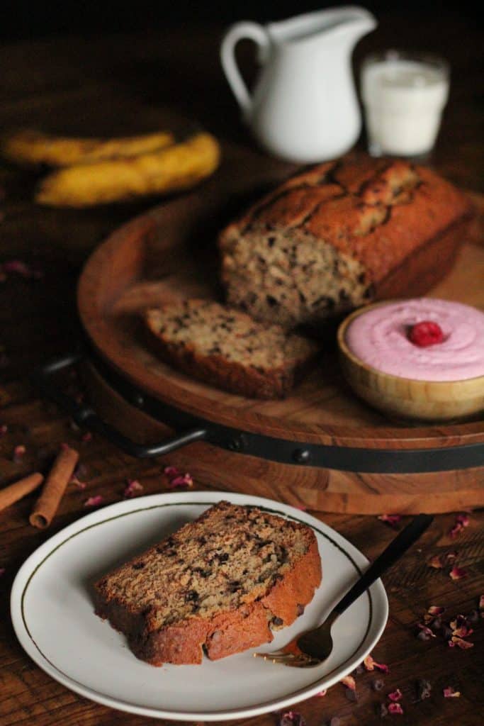 slice of banana bread on white plate and loaf of chocolate chip banana bread and a bowl of raspberry mascarpone cream in background