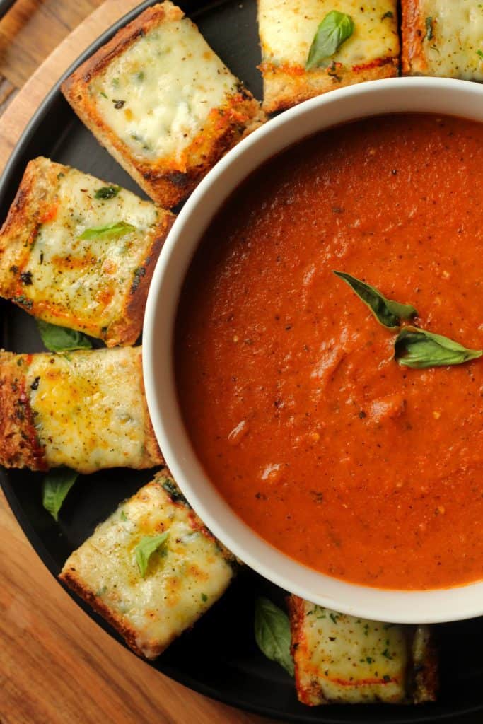 roasted tomato basil and red pepper soup in a white bowl with slices of cheesy garlic bread on a black plate on a brown surface