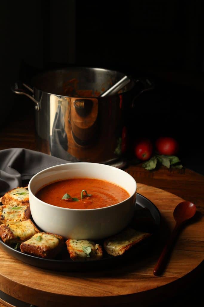 roasted tomato basil and red pepper soup in a white bowl with slices of cheesy garlic bread on a black plate on a brown surface3