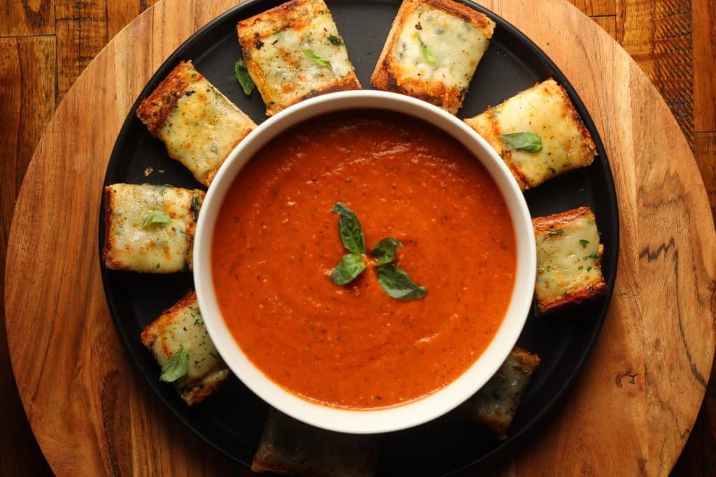 roasted tomato basil soup in a white bowl with slices of cheesy garlic bread on a black plate on a brown surface