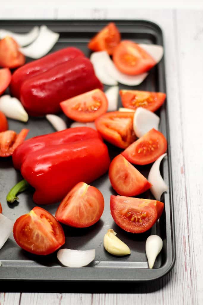 sheet pan with raw sliced onions and tomatoes, halved red bell peppers, and peeled garlic cloves