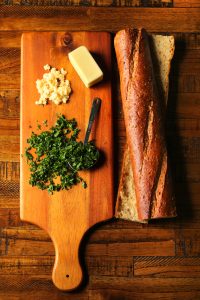 sliced multigrain baguette on a brown wooden surface with minced garlic, chopped parsley, and butter on a wooden cutting board