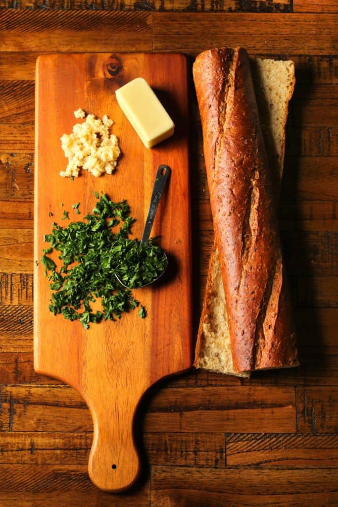 sliced multigrain baguette on a brown wooden surface with minced garlic, chopped parsley, and butter on a wooden cutting board