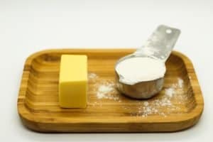 butter and a stainless steel measuring cup filled with flour plus more scattered on a light brown wooden plate on a white surface