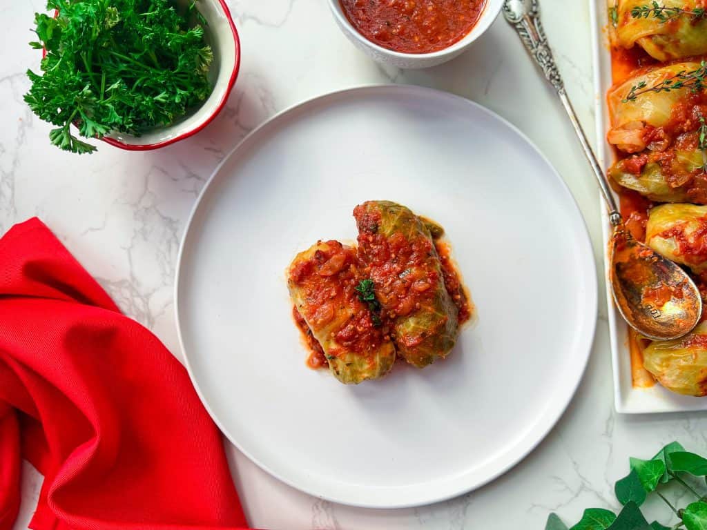 two stuffed cabbage rolls topped with tomato sauce on a white plate, bright red napkin, white serving plate with stuffed cabbage rolls, extra sauce in a bowl, and a red bowl filled with parsley