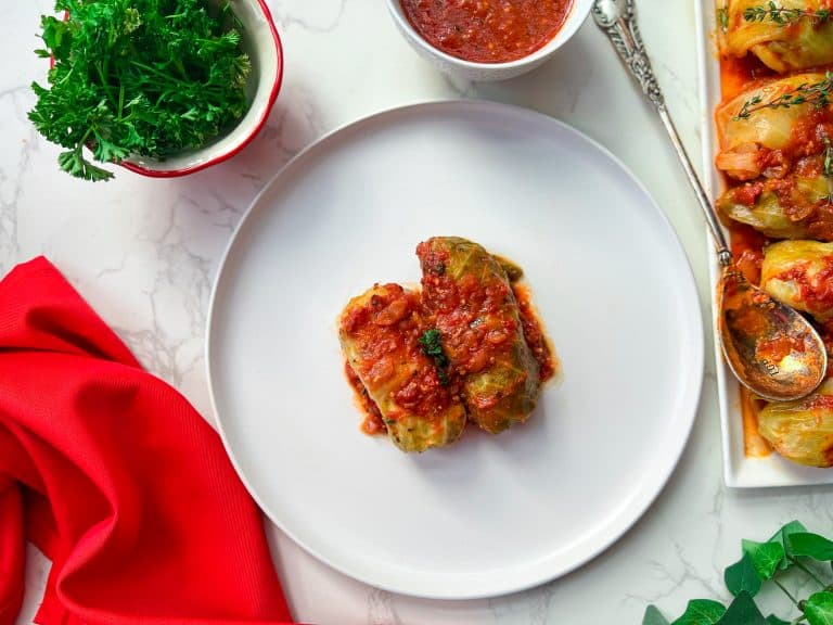 two stuffed cabbage rolls topped with tomato sauce on a white plate, bright red napkin, white serving plate with stuffed cabbage rolls, extra sauce in a bowl, and a red bowl filled with parsley