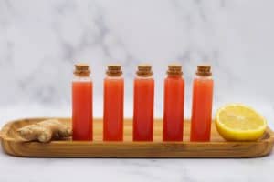 5 watermelon lemon ginger juice wellness shots in tiny glass vials on a light wooden cutting board.