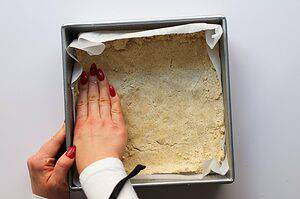 Shortbread pie crust being pressed into a cake pan lined with parchment paper.