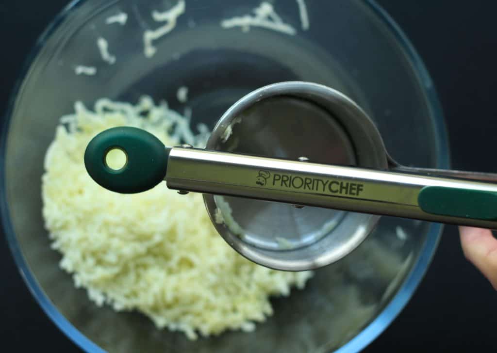 A Piority Chef brand ricer ricing potatoes into a glass bowl on a black surface.