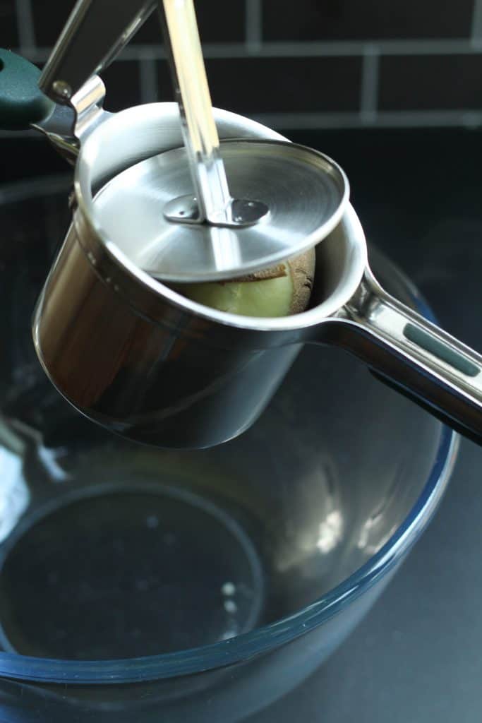 A potato in a ricer over a glass mixing bowl.