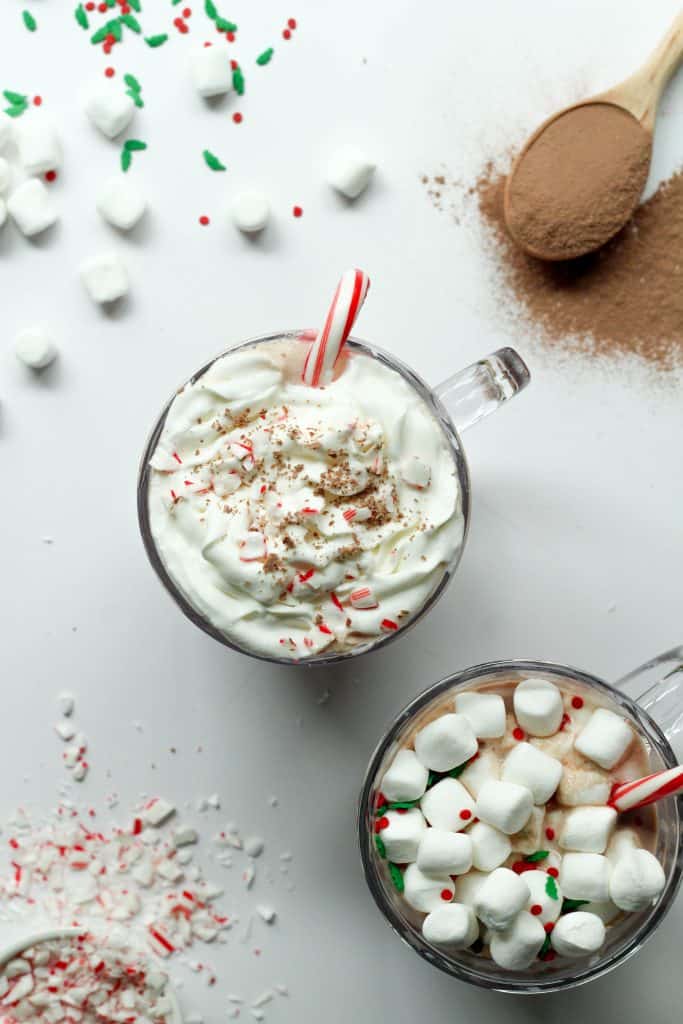 Two cups of protein hot chocolate. One with whipped cream and a peppermint candy cane, and one with marshmallows.