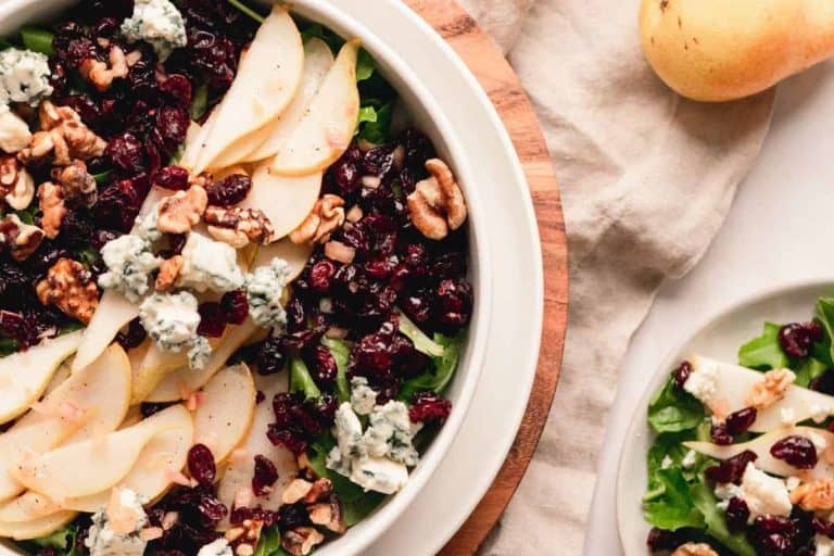 Pear blue cheese salad topped with dried cranberries in a white bowl with a beige napkin and whole pear in the photo.