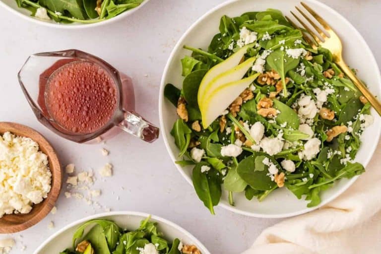 Walnut pear salad on three white plates with a gold fork and pink dressing in a clear class serving cup with a handle.