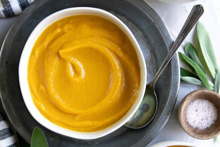 Swirled acorn squash soup in a white bowl on a gray plate with a silver spoon and a small wooden bowl of salt and sage leaves.