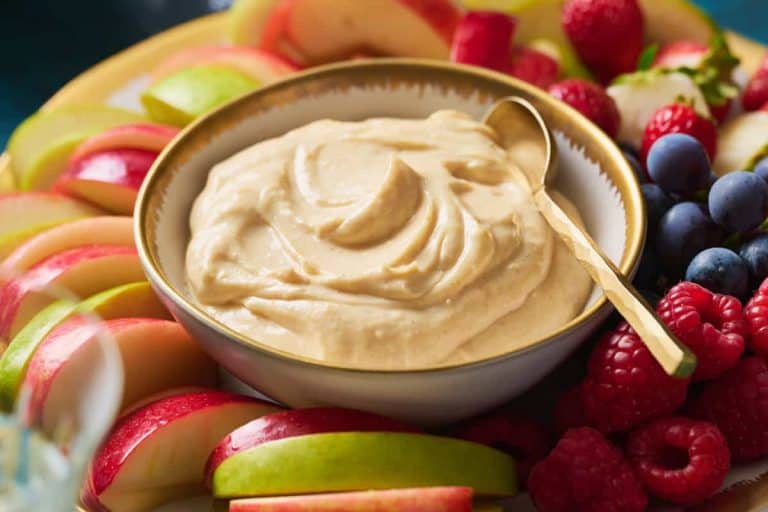 Cheesecake fruit dip recipe swirled in a gold rimmed white bowl with a gold spoon surrounded by berries and apple slices.