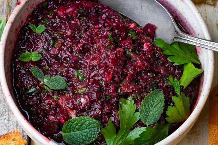 Beautoful cranberry ezme relish in a white speckled bowl topped with fresh green parsley and mint leaves along the side of bowl.