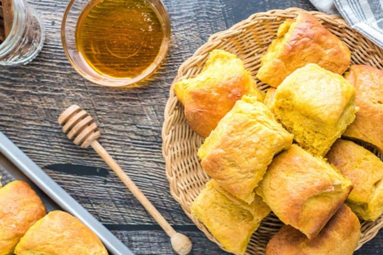 Honey pumpkin dinner rolls piled in a wicker basket on a brown wooden surface with a honey drizzling