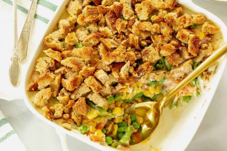 Mixed vegetable casserole topped with crusty bread pieces in a white dish with a cold spoon.