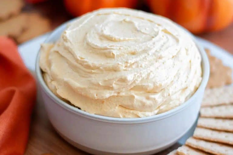 Pumkin cool whip dip in a white bowl with an orange napkin and orange pumpkins blurred in the background.