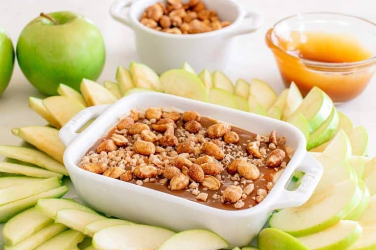 Caramel apple dip topped with nuts and caramel in a white baking dish with handles surrounded by sliced green apples.