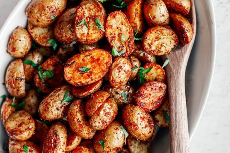 Crispy roasted mini potatoes sprinkled with chopped fresh parsley piled in a white oval dish with a wooden spoon.