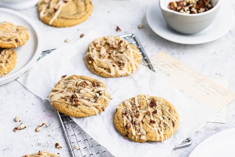 Three maple cookies drizzled with icing and topped with pecans on a silver wire rack topped with parchment paper.