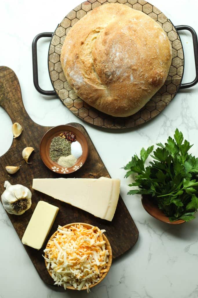 Stuffed garlic bread ingredients (cheese, butter, parsley, garlic, bread loaf seasonings) displayed on wooden boards.