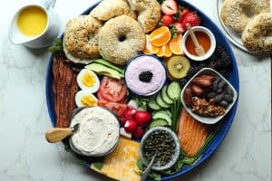 Bagel board charcuterie style with sweet and savory spreads, fruit, vegetables, salmon lox, bacon, eggs, and a pitcher of orange juice.