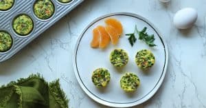 Cooked veggie egg bites on a white plate with orange slices and more egg bites in the mini muffin pan.