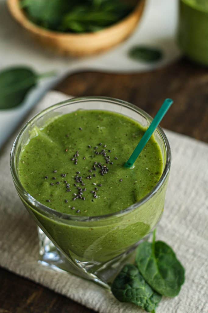 Green protein smoothie with a turquoise-colored straw with spinach leaves on a cloth lined wooden board.