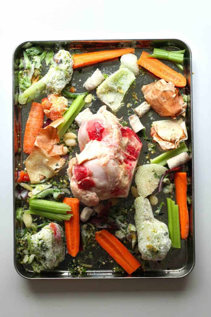 A raw beef bone on a sheet pan with scattered veggie scraps for homemade bone broth.