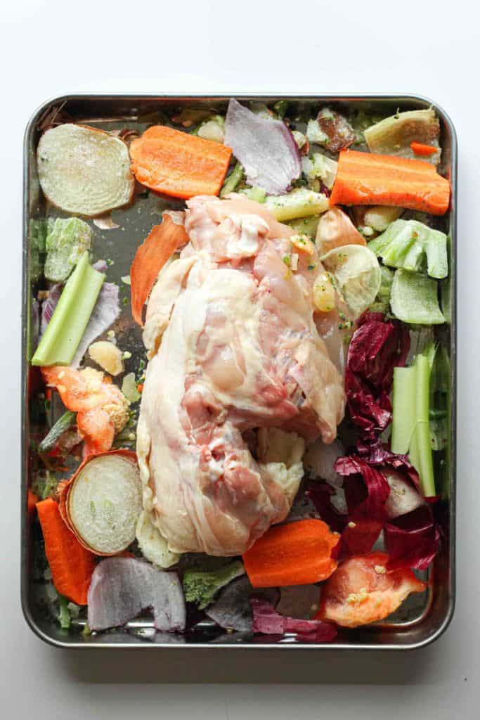 A raw chicken carcass on a sheet pan with veggie scraps and vegetables like carrots, onion, and celery.
