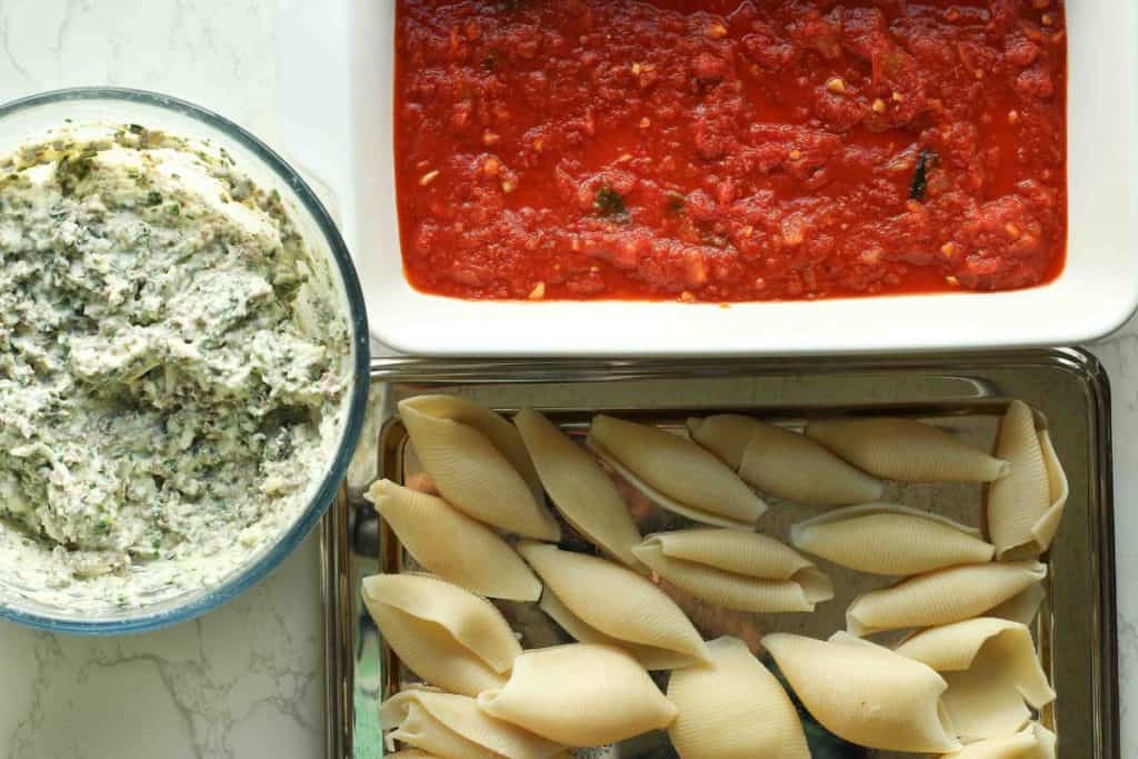A bowl with cheese and beef mixture, a tray of cooked jumbo pasta shells, and a baking dish with a layer of marinara sauce in the bottom.
