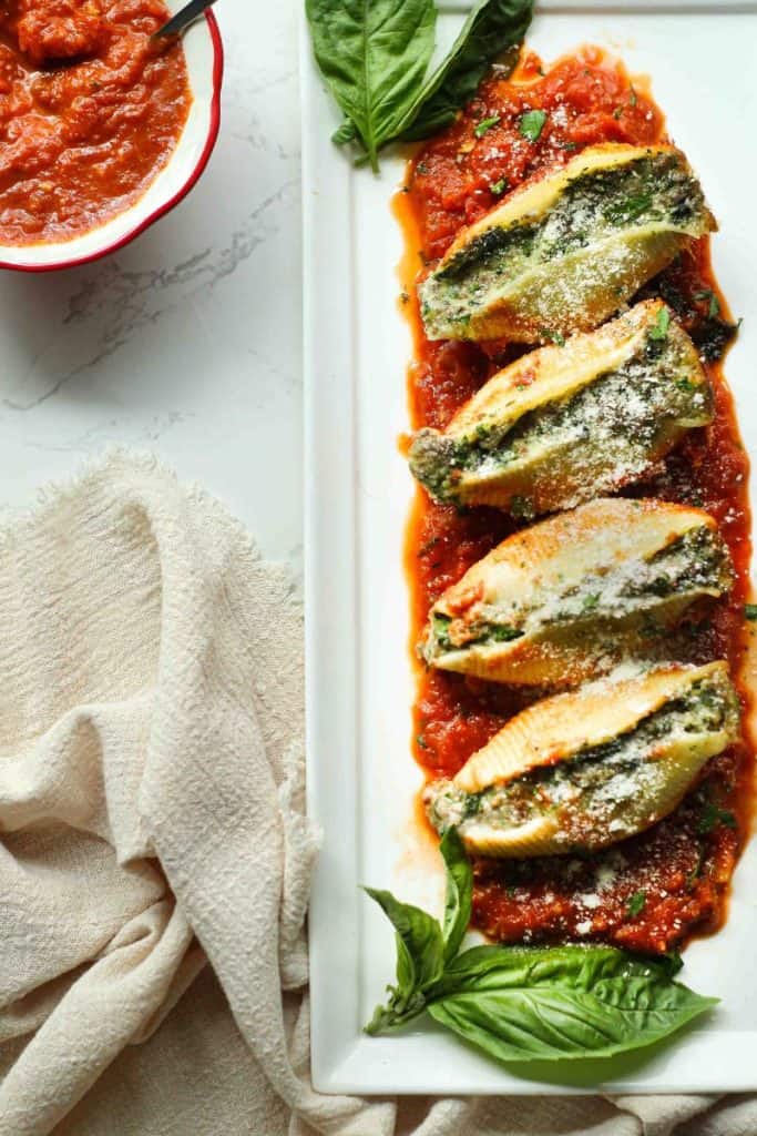 4 beef stuffed shells line on a bed of sauce on a white plate with whole basil leaves with a beige napkin and side of sauce in red bowl.