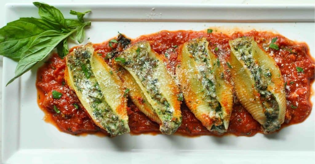 Four beef stuffed shells sprinkled with parmesan on a bed of chunky marinara sauce on a white plate with a sprig of basil.