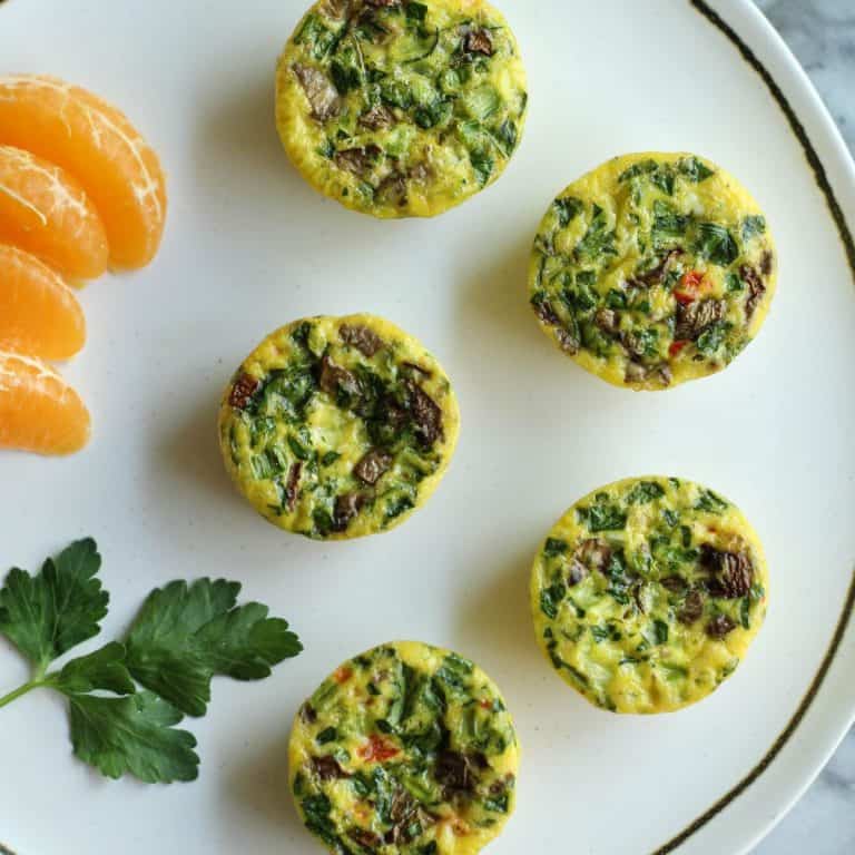 Five veggie egg bites displayed on a round white plate with orange slices and orange juice and green napkin in background.