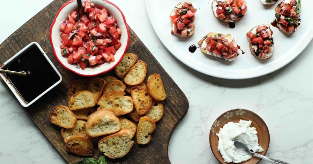 Tomato bruschetta on crostini with balsamic glaze on a plate and a dish of goat cheese, plus more crostini on a cutting board.