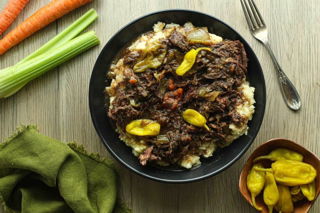 Instant pot Mississippi pot roast with pepperoncinis over mashed potatoes in a black bowl with carrots and celery stalks in corner.