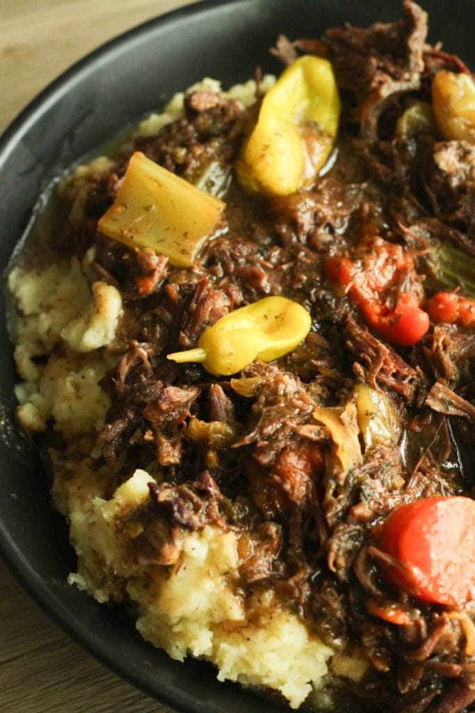 Instant pot mississippi pot roast over mashed potatoes in a black bowl topped with whole pepperoncini and diced carrots in a black bowl.