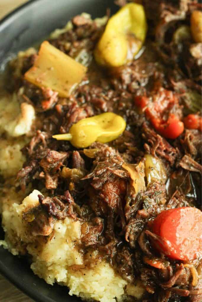 Instant pot mississippi pot roast over mashed potatoes in a black bowl topped with whole pepperoncini and diced carrots in a black bowl.
